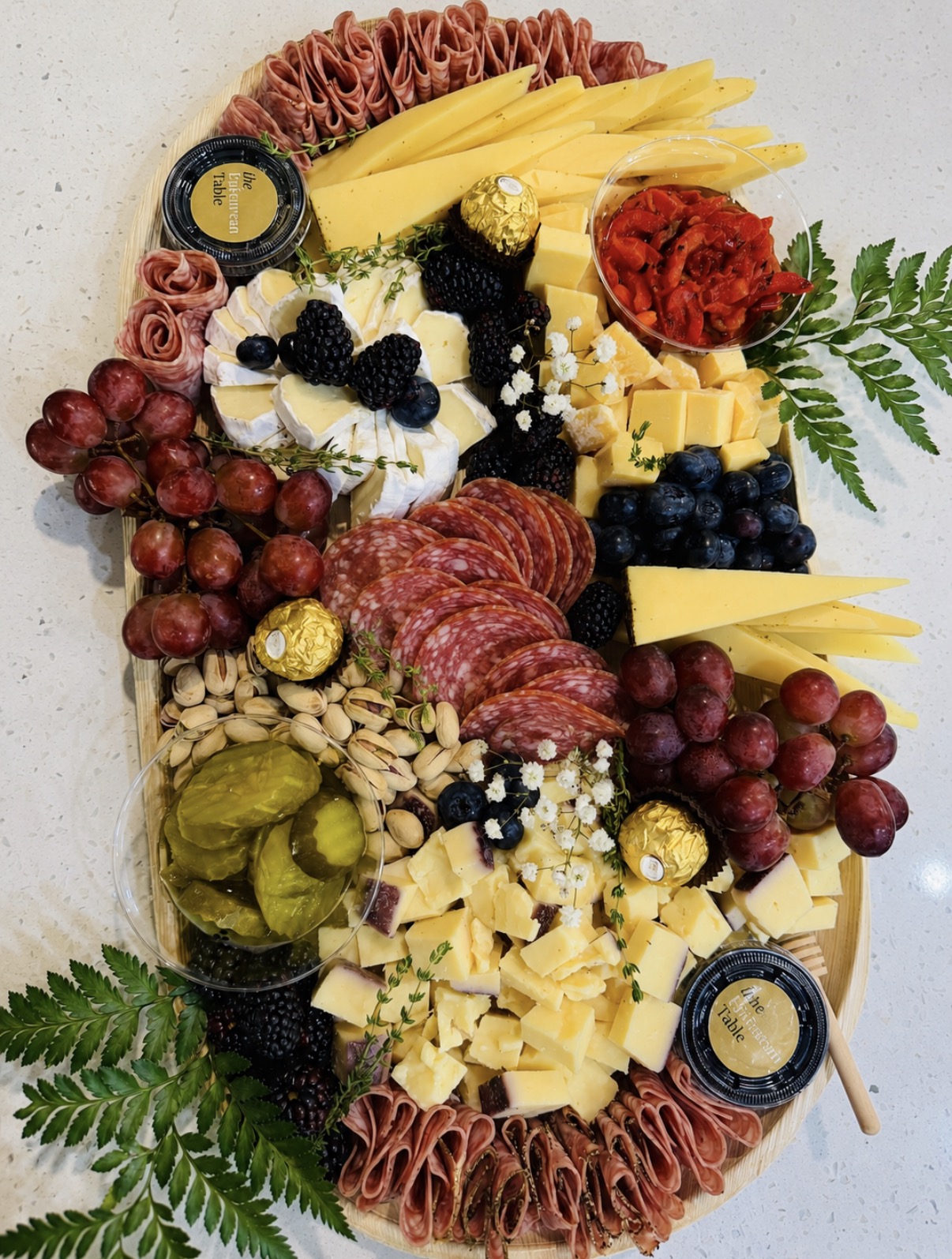 The Epicurean Table board with cheese fans, red grapes, salami, berries, and greenery